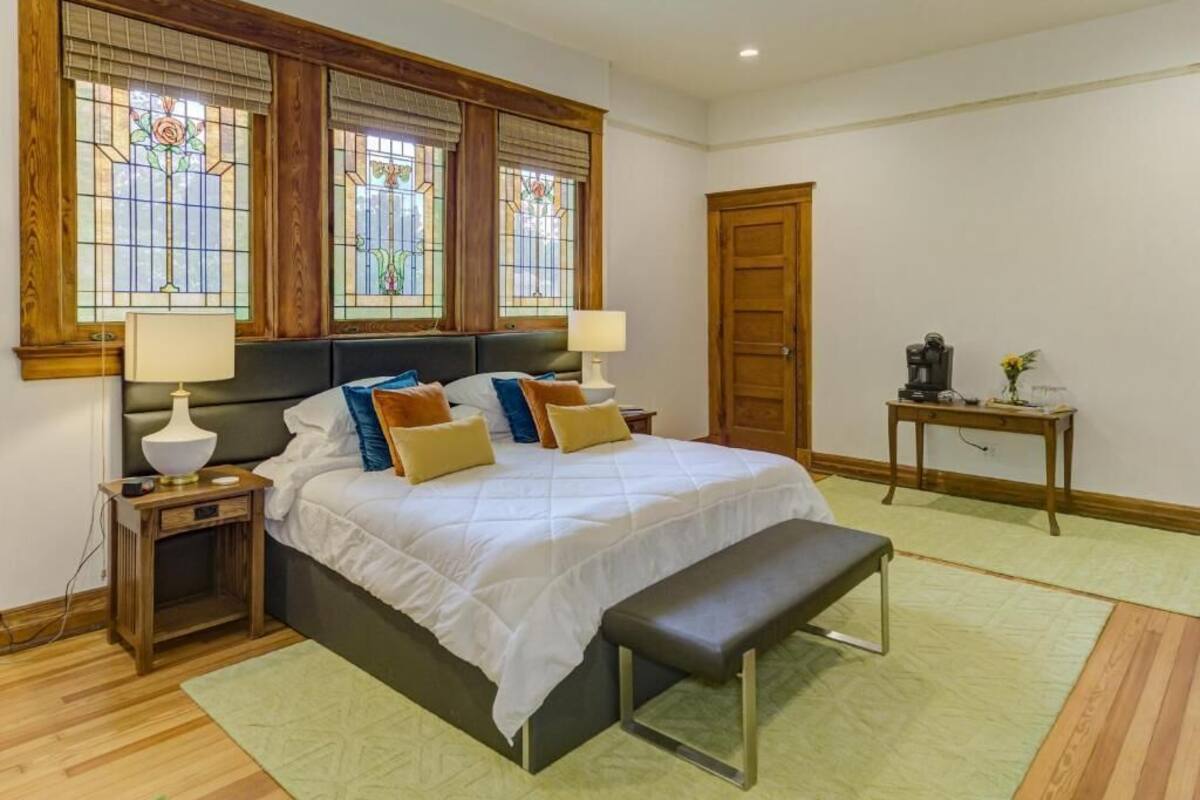 A bedroom with a large bed, two bedside tables with lamps, a bench, a wooden door, and a small table with a vase.