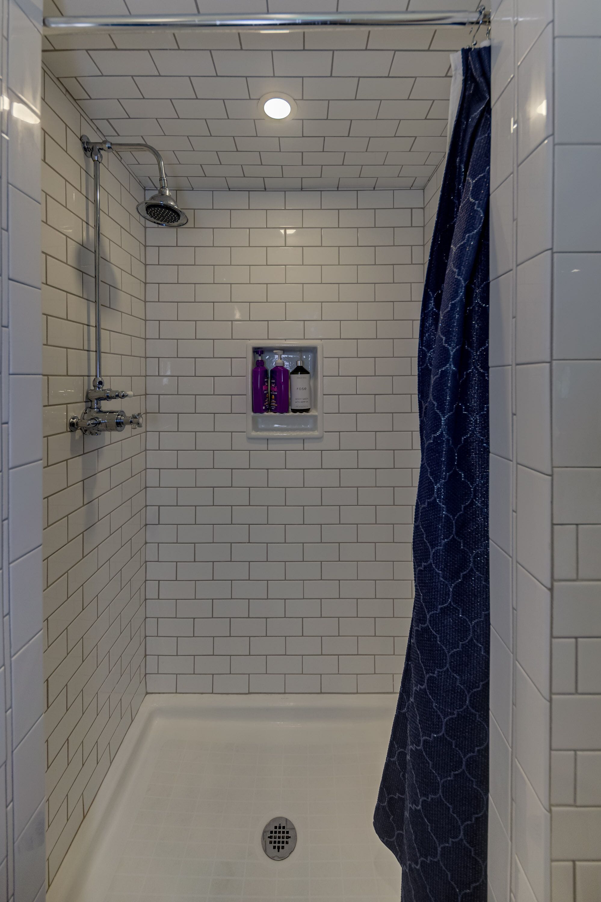 A walk-in shower with white tiled walls, a showerhead, a handheld shower attachment, a blue shower curtain, and a shelf with two bottles.
