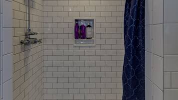 A walk-in shower with white tiled walls, a showerhead, a handheld shower attachment, a blue shower curtain, and a shelf with two bottles.