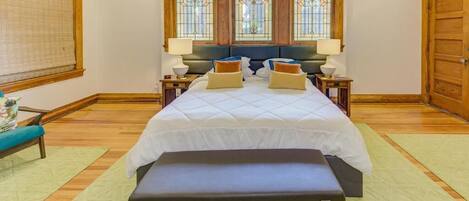 Un dormitorio con una cama grande, cabecera de madera, dos mesitas de noche con lámparas, un banco al pie de la cama y una ventana con vidrieras.