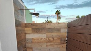 A narrow outdoor shower area with a wooden wall and floor, a metal showerhead, and a view of palm trees and the sky.