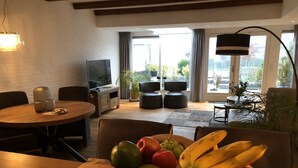 A modern living room with a wooden dining table, a fruit bowl, a TV, and a large window with curtains.