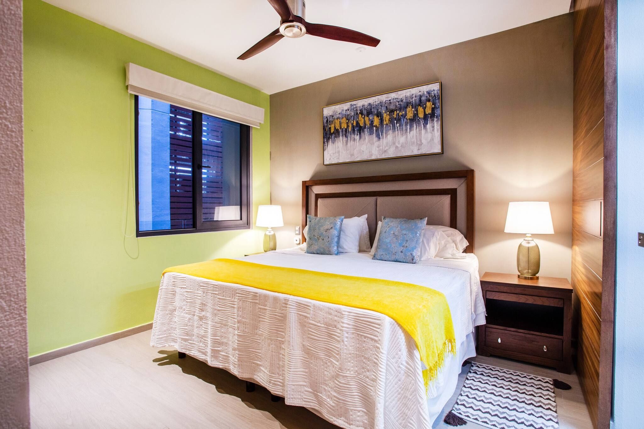 A bedroom with a bed, two bedside lamps, a ceiling fan, a window with blinds, and a painting on the wall.