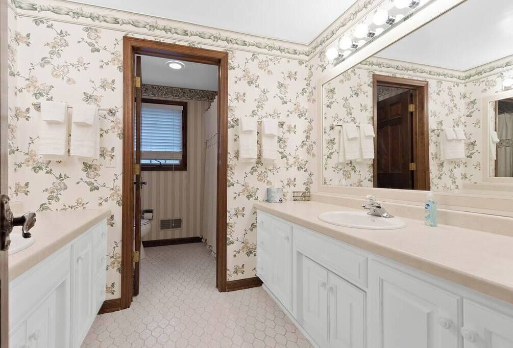 Een badkamer met bloemen behang, een grote spiegel, een wastafel en een deur naar een andere kamer.