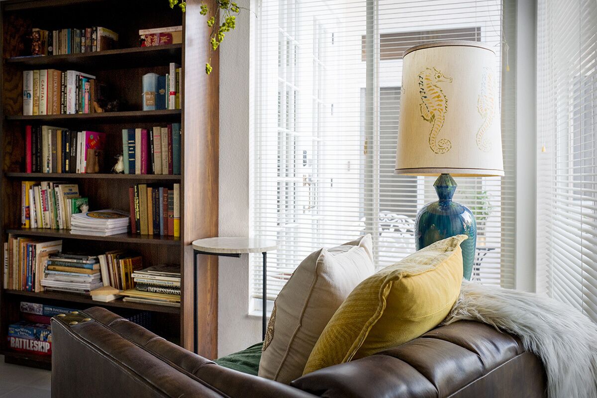 A cozy living room with a leather sofa, a bookshelf filled with books, a lamp with a seahorse design, and a window with blinds.