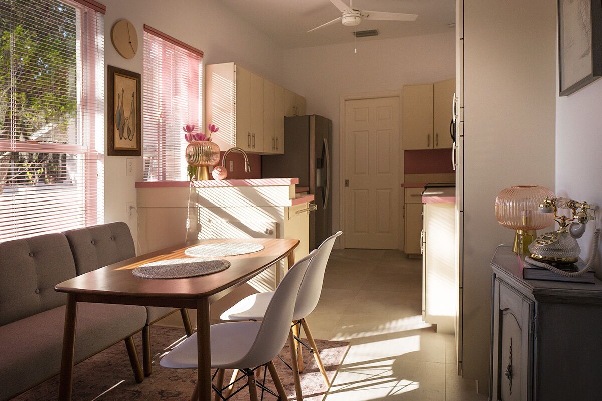 A dining area with a wooden table, white chairs, and a sofa. There is a kitchen in the background with cabinets and a refrigerator.
