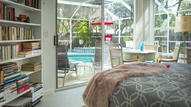 A room with a large glass door leading to a pool area, a bookshelf filled with books, a bed with a patterned cover, and a small table with a lamp. - :: Villa Oasis – Elegant & Peaceful Pool Retreat in Siesta Key :: (Siesta Key)