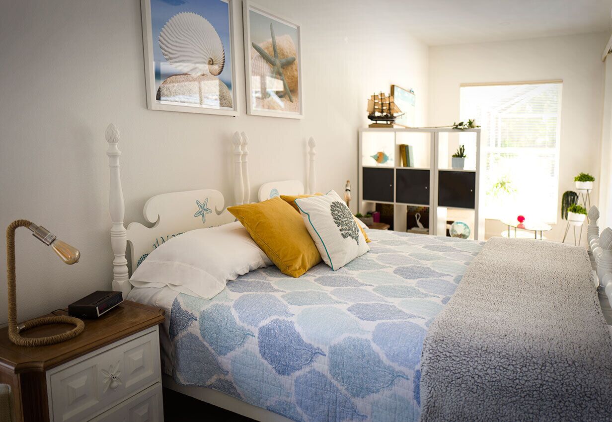A bedroom with a bed, a nightstand, a lamp, a bookshelf, and a window.