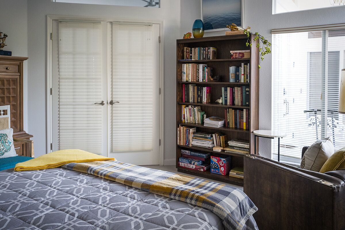 Une chambre à coucher avec un lit, une bibliothèque, une fenêtre avec des stores et une porte.