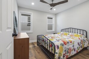 A bedroom with a bed, a wooden dresser, two windows with blinds, a ceiling fan, and a white door.