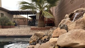 Um jardim rochoso com uma pequena cascata, uma palmeira e um edifício com fachada de tijolos ao fundo.