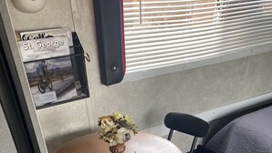 Uma área de refeições compacta com uma mesa redonda, duas cadeiras pretas, um vaso com flores, um suporte para revistas e uma janela com persianas.