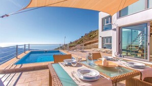 Un área de piscina en la azotea con vistas despejadas a la costa, un espacio de comedor con mesa y sillas, y un edificio moderno con grandes ventanales.