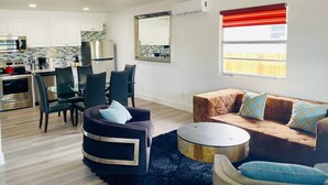 Un salon moderne comprenant un coin repas, une cuisine avec des meubles blancs et une grande fenêtre agrémentée de stores rouges.