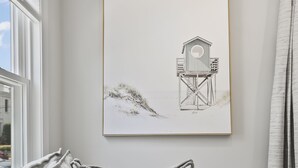 Habitación en tonos neutros con un sofá gris, una obra enmarcada que representa una estructura de madera y una ventana con cortinas.