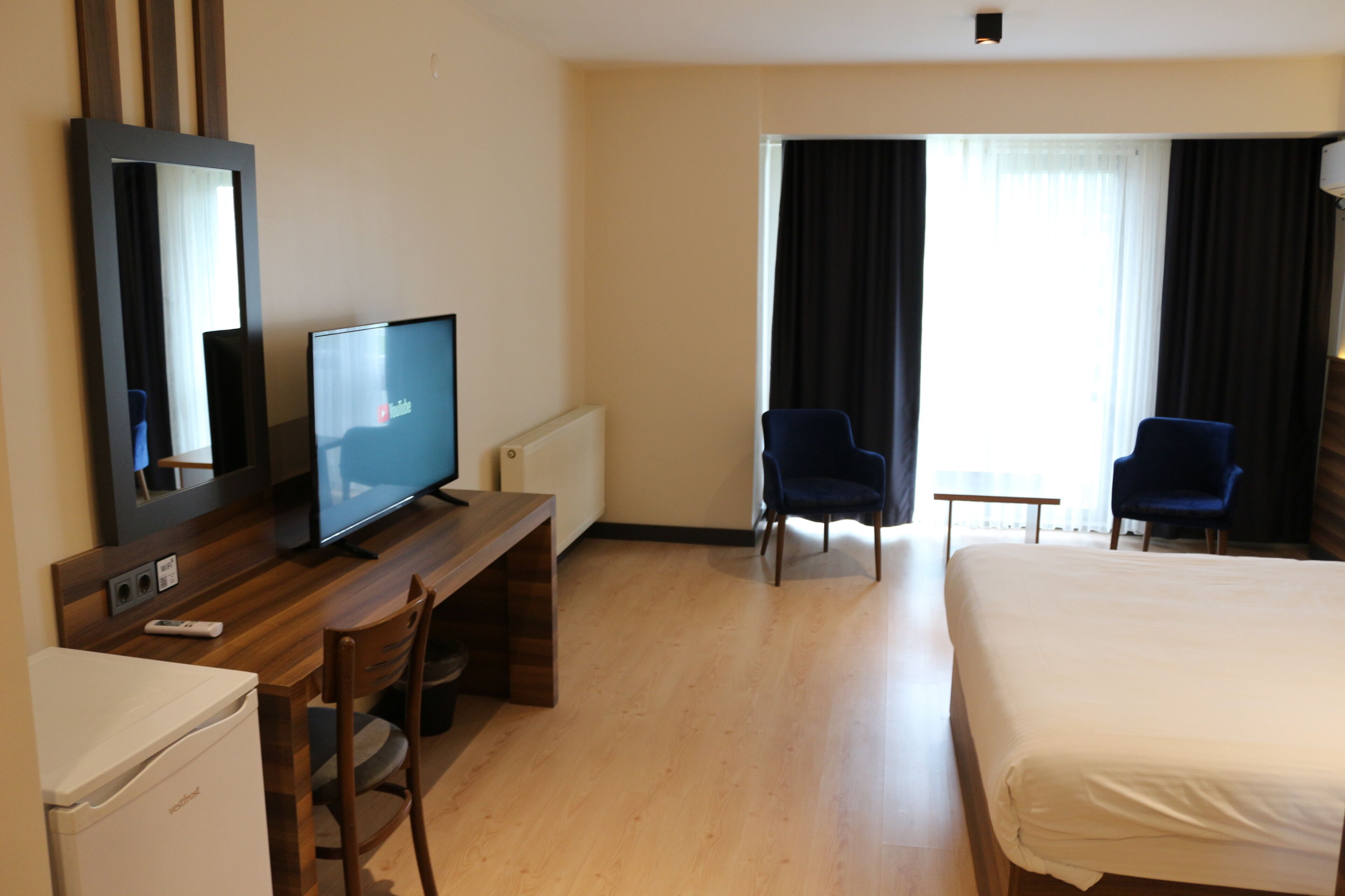 Habitación de hotel con cama, escritorio con televisor, dos sillas, una mesita y ventana con cortinas.