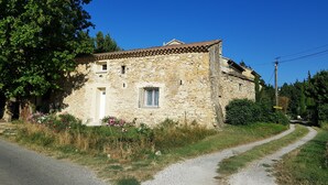 A stone building with a tiled roof, a small window with a metal grille, and a door. There is a gravel path leading to the building, surrounded by greenery and a clear blue sky. - Quiet house in the countryside (CADEROUSSE)