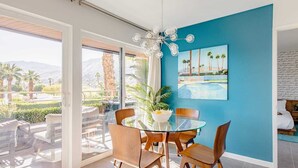 Un comedor moderno con una mesa de vidrio y sillas de madera, una pared azul como detalle y vistas a palmeras y montañas.