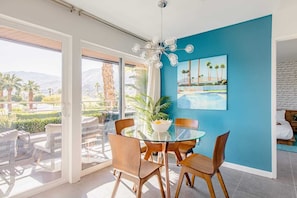 Un comedor moderno con una mesa de vidrio y sillas de madera, una pared azul como detalle y vistas a palmeras y montañas.
