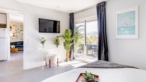 Un dormitorio moderno con una cama grande, un televisor montado en la pared, un balcón con vistas panorámicas y una zona de comedor visible a través de una puerta abierta.
