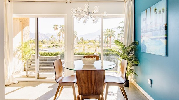 Un comedor con mesa y sillas de vidrio, una araña y vistas a palmeras y montañas.