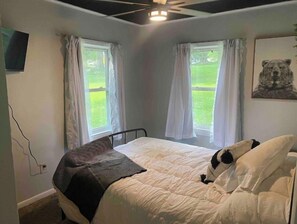 Une chambre à coucher avec un lit, deux fenêtres avec des rideaux, un ventilateur de plafond, un téléviseur fixé au mur et un tableau représentant un ours.