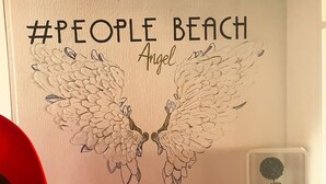 Een muur met een #PEOPLE BEACH Angel ontwerp, een rode stoel en een wandplank met potplanten.