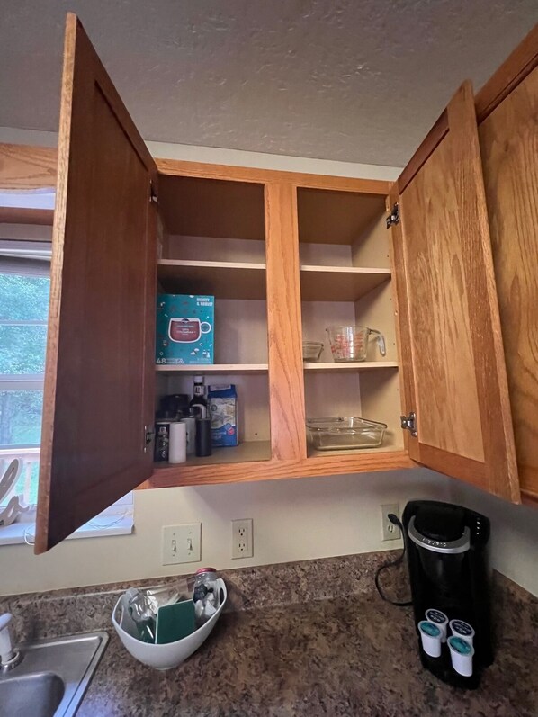 A kitchen with wooden cabinets, a coffee maker, a bowl with utensils, and a window with a view of trees.