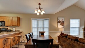 Un comedor con una mesa de madera, cuatro taburetes altos y un banco. Hay una zona de cocina con armarios de madera y un microondas. Un candelabro cuelga del techo, y hay dos ventanas con persianas.