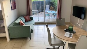 A room with a green sofa, a dining table with chairs, a TV mounted on the wall, and a sliding glass door leading to an outdoor area with a table and chairs. - Apartment between lake and sea amidst vineyards (Peret)