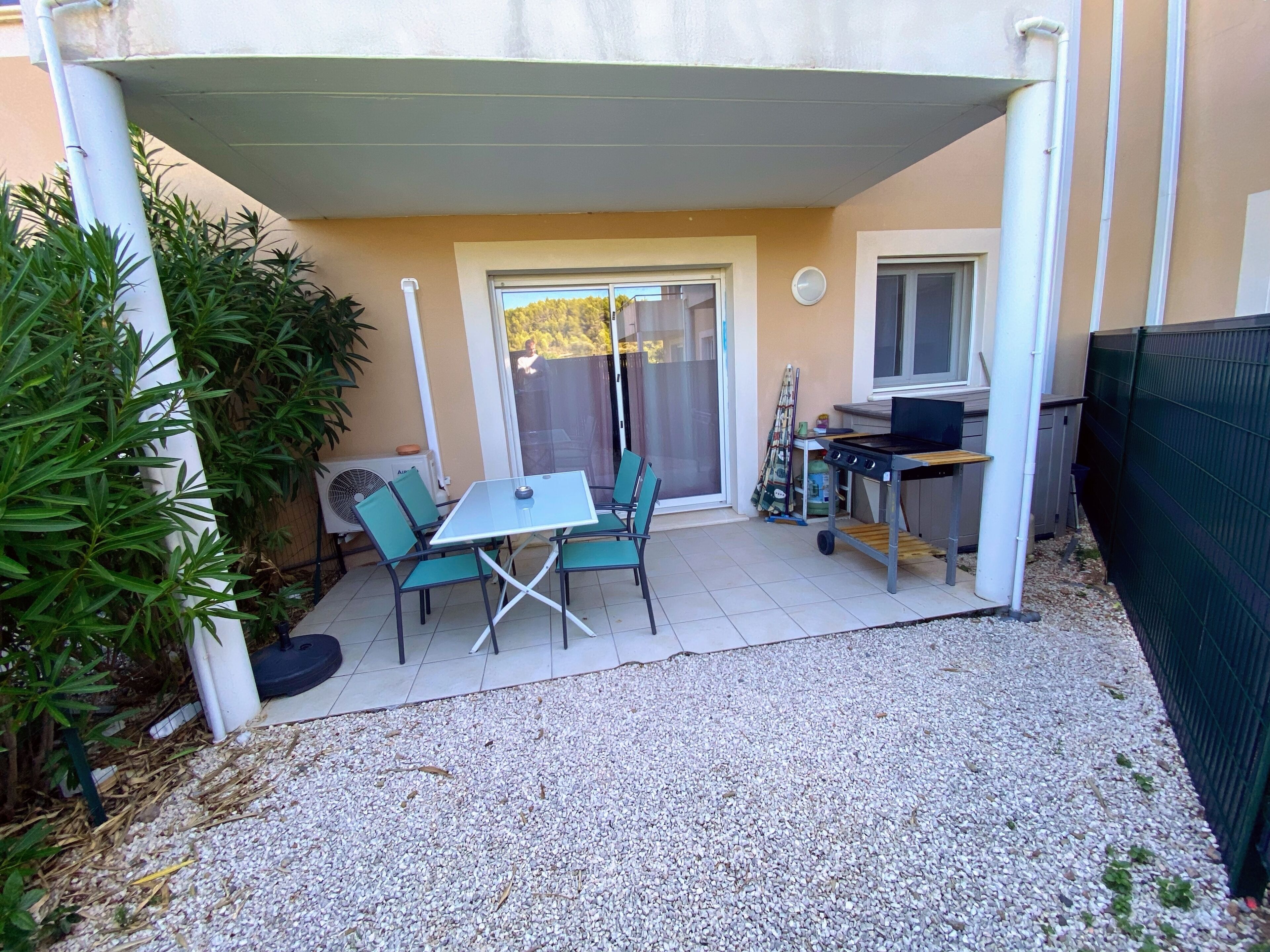 A covered patio area with a table and chairs, a grill, and a window overlooking a garden.