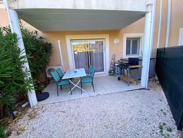 Uma varanda coberta com mesa, cadeiras, churrasqueira e janela com vista para um jardim.