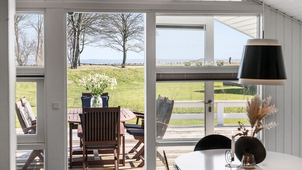 Een kamer met een groot raam dat uitkijkt op een grasveld en een waterpartij. Er staat een tafel met stoelen en een bloevas.
