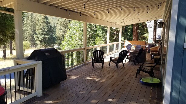 Een overdekt terras buiten met houten vloer, zwarte Adirondack-stoelen en een barbecue.