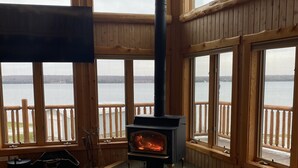 Una habitación acogedora con estufa de leña, ventanas amplias con vista a un lago, suelo de madera y una mesa.