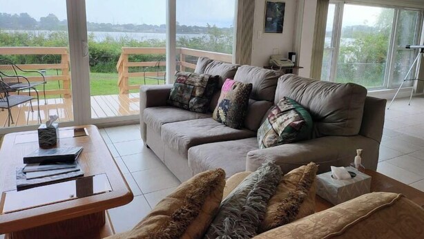 Amplia sala de estar con un gran ventanal que ofrece vistas a un lago y follaje. Hay un sofá con cojines estampados y una mesa de centro de madera con libros y un florero.