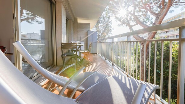 A balcony with a view of a tree and buildings, a table with chairs, and a refrigerator. - Renovated 3-Room Apartment in a Leafy Park (Cannes)