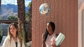 Two people holding rolled-up mats, a camera, and a disco ball in a covered outdoor area with palm trees and mountains in the background. - Mr. Pink's MCM Alexander with Pool, Hot Tub, Fire Pit (Palm Springs)