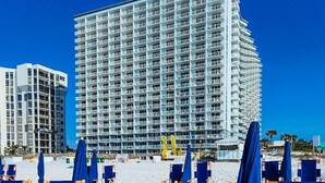 Un hotel frente a la playa con varios pisos, una zona de playa arenosa con sillas azules tipo lounge, y un cielo azul despejado.