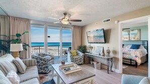 Amplia sala de estar con un ventanal con vista a la playa, ventilador de techo y cama con colcha estampada.