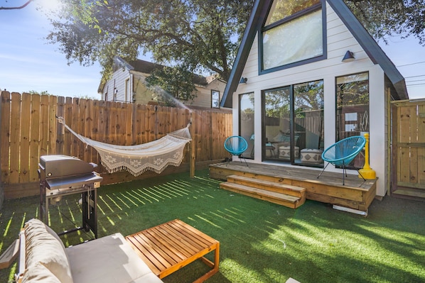 A backyard with a hammock, a grill, and a wooden deck leading to a house with large windows.