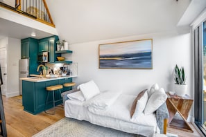 A modern kitchen with teal cabinets, a central island, and bar stools. There is a white sofa with a view of the sea through a large window.