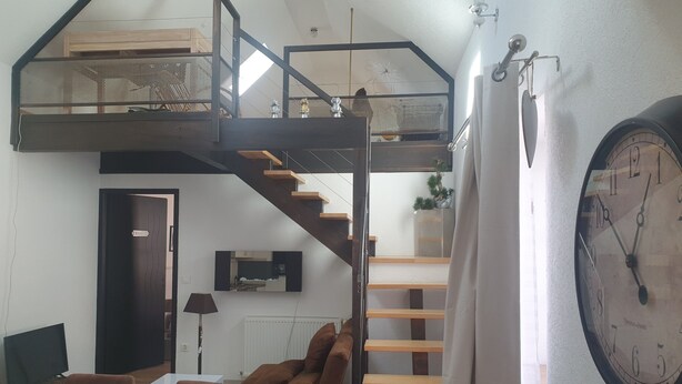 Un espace de vie moderne de style loft, doté d’un escalier menant à un niveau supérieur, d’un coin repas avec une table et des chaises, et d’une grande horloge murale.