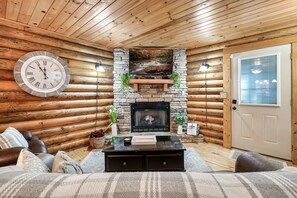 A log cabin interior with a stone fireplace, a clock, and a cozy seating area.