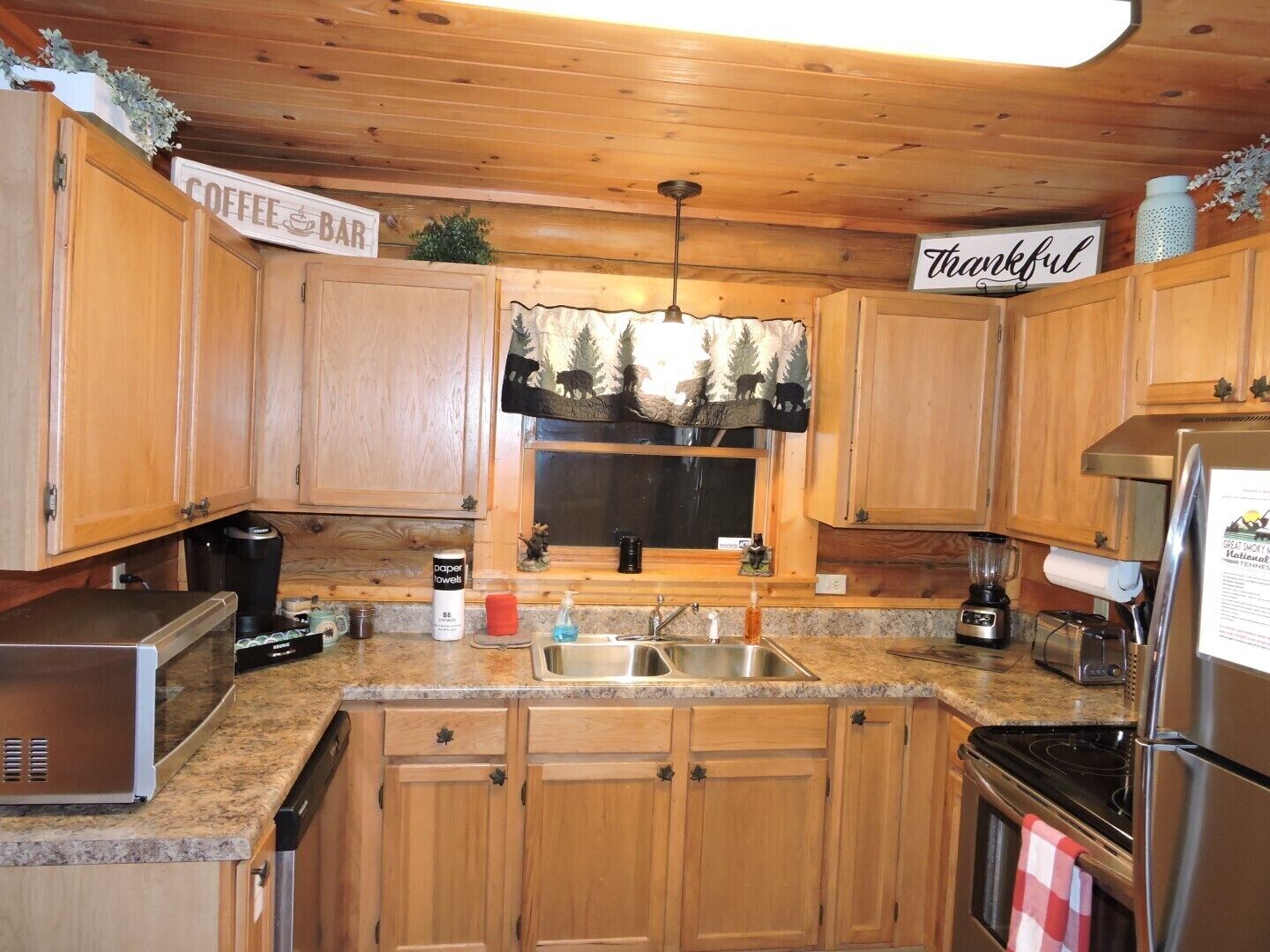 Una cocina con armarios de madera, un letrero que dice 'Coffee Bar', una ventana con cortina de motivos forestales y un letrero con la palabra 'Thankful'.