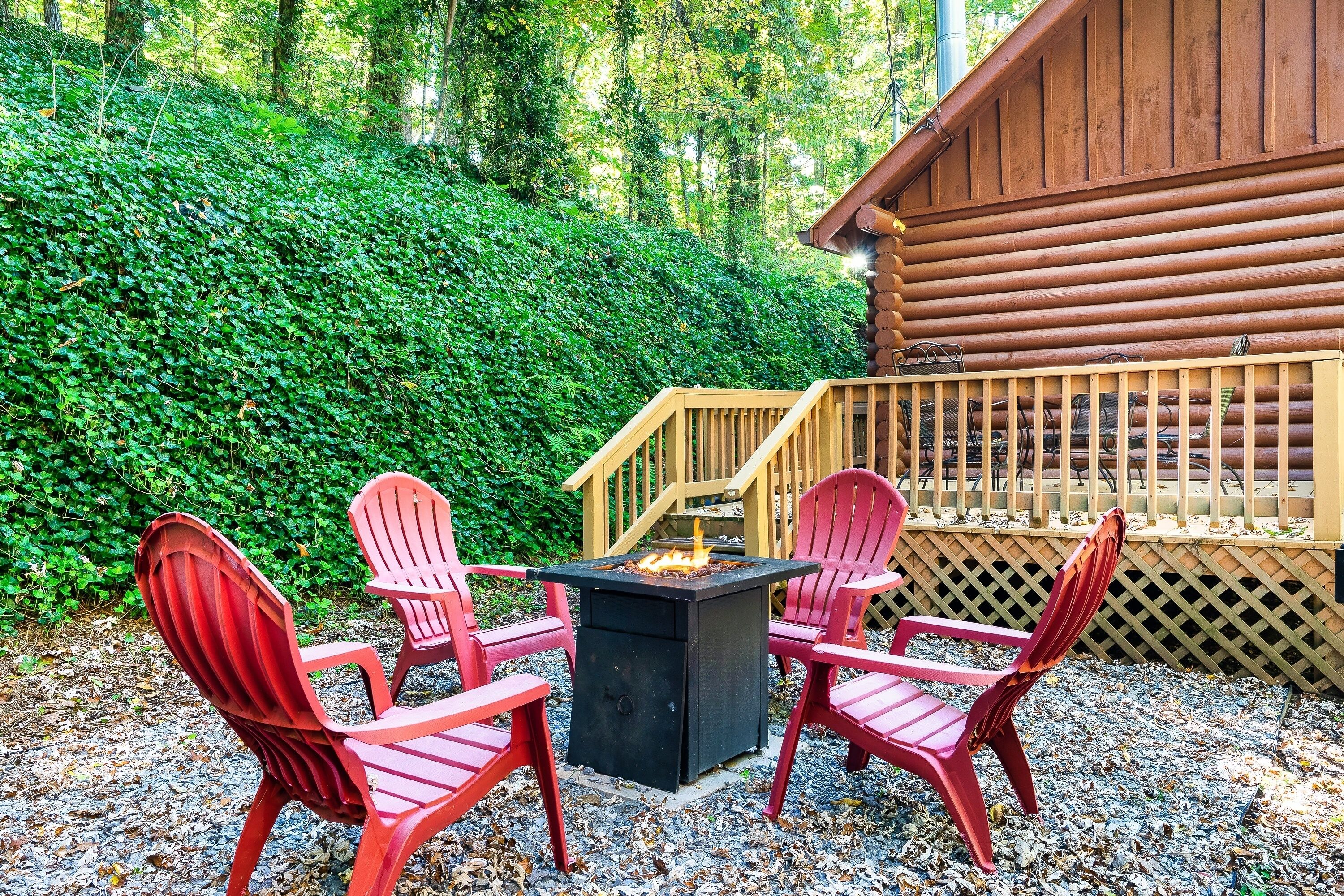 Un patio con cuatro sillas Adirondack rojas alrededor de un brasero cuadrado, una terraza de madera y una cabaña al fondo.