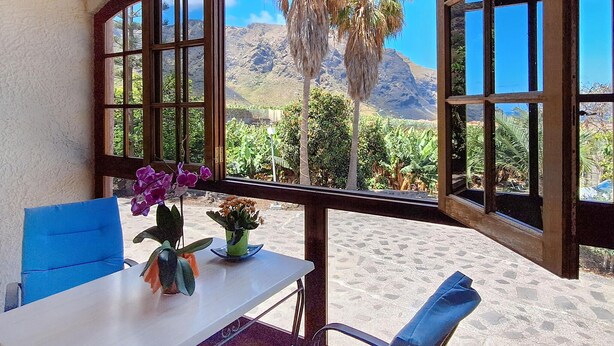 A room with a table and chairs, a window with a view of a mountainous landscape and palm trees. - 1 bedroom amazing apartment in Buenavista Del Norte (Buenavista Del Norte)