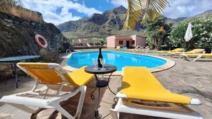 A pool area with lounge chairs and a table, surrounded by mountains and greenery. - 1 bedroom amazing apartment in Buenavista Del Norte (Buenavista Del Norte)
