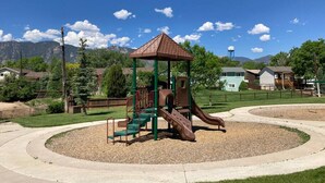 A playground with a slide and climbing structures, surrounded by a landscaped area with a curved path. - 5BR Recently Updated Retreat w/ Hot Tub, BBQ grill, Fire Pit, Near Attractions (Colorado Springs)