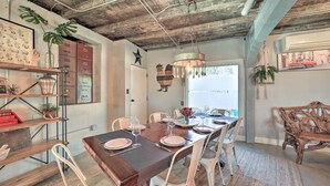 A dining area with a wooden table set for six, surrounded by white chairs. A chandelier hangs above, and there are plants on shelves and a window with a view. - Eclectic Asheville Abode w/ Backyard Oasis! (Asheville)